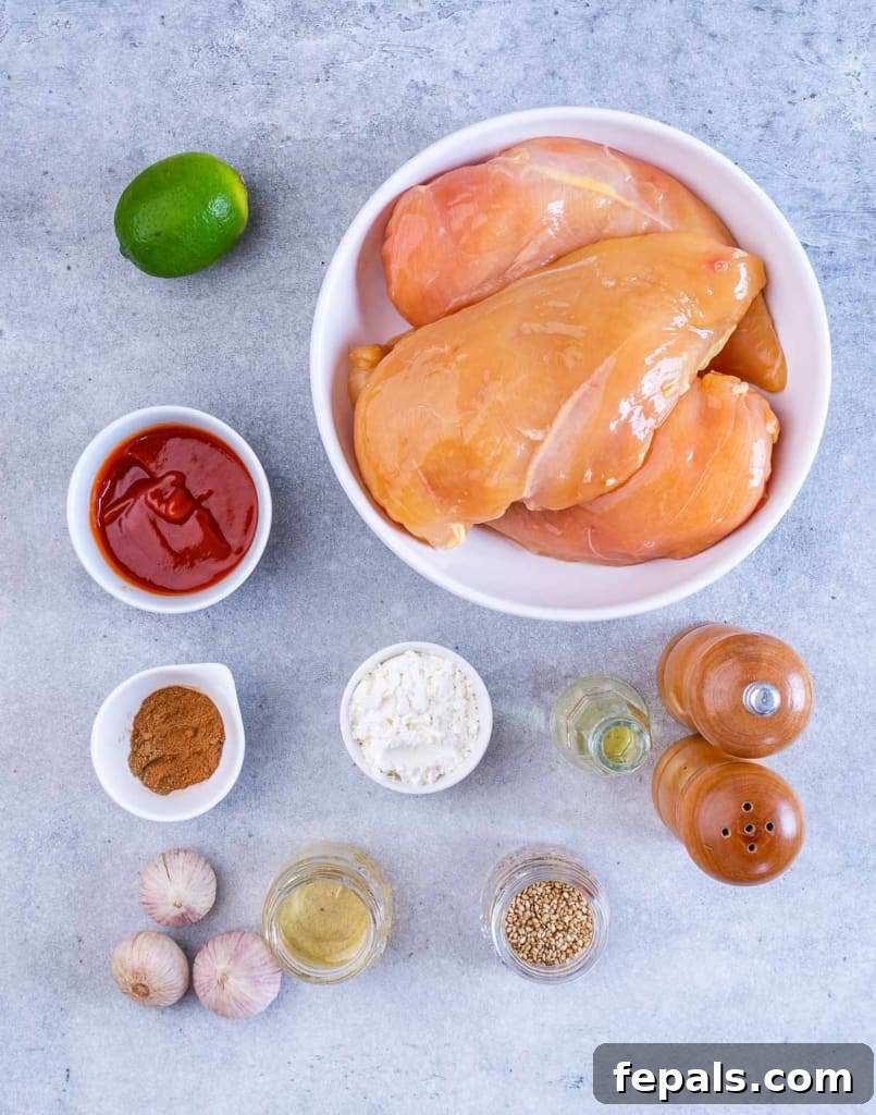 Sticky Honey Sriracha Chicken 4 A collection of ingredients laid out on a table, including chicken breasts, sriracha bottle, honey, lime, garlic, spices, and sesame seeds, ready to be used for baking honey sriracha chicken.