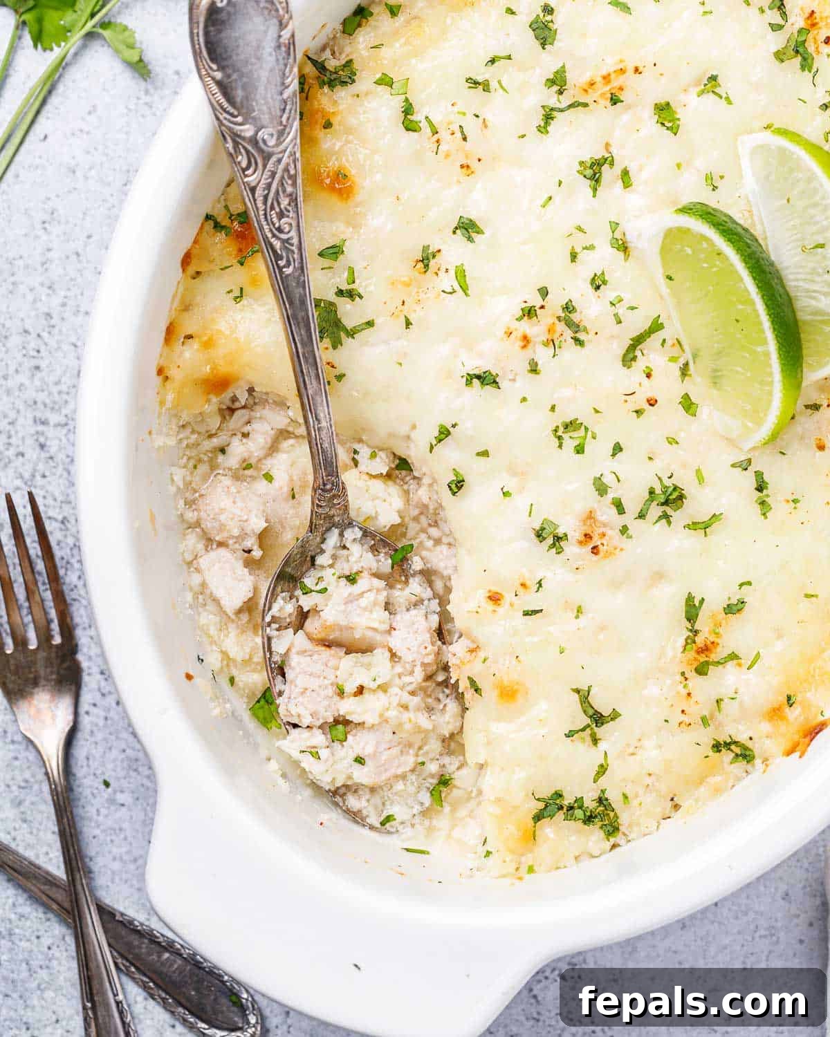 A spoon lifting a generous portion of cheesy chicken and cauliflower rice casserole from a white baking dish, showing the creamy texture and melted cheese.