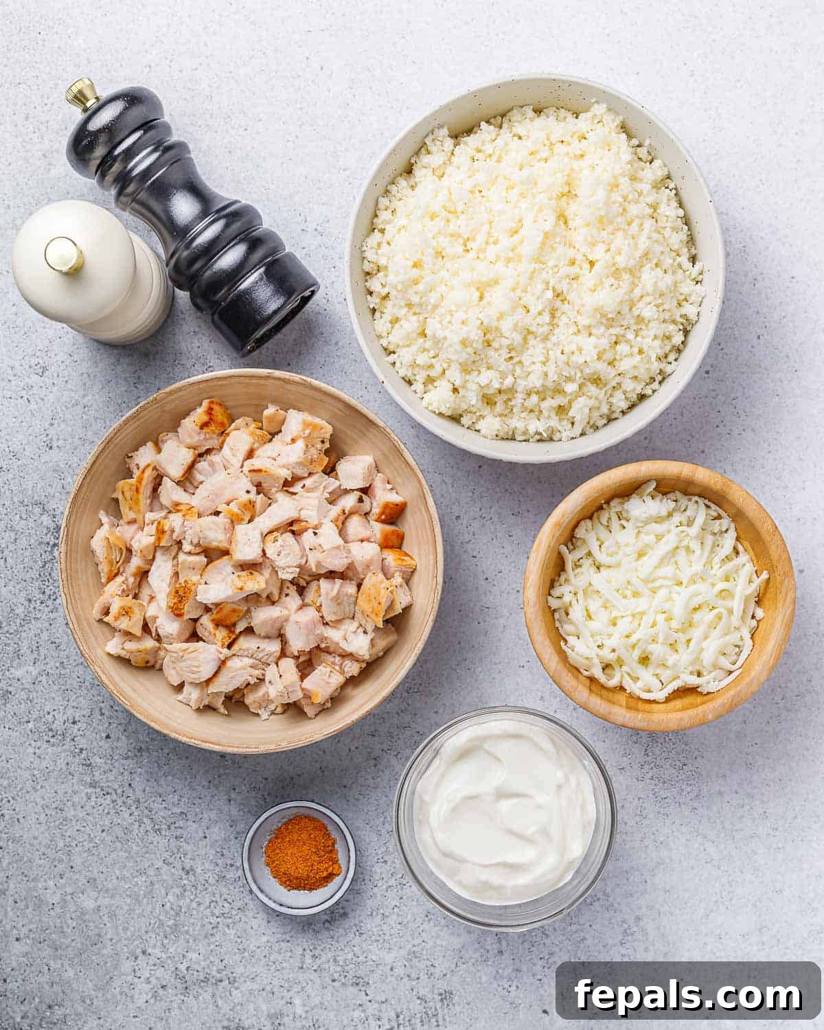 Small bowls displaying separated ingredients: chopped chicken, cauliflower rice, shredded cheese, and sour cream, ready for mixing.