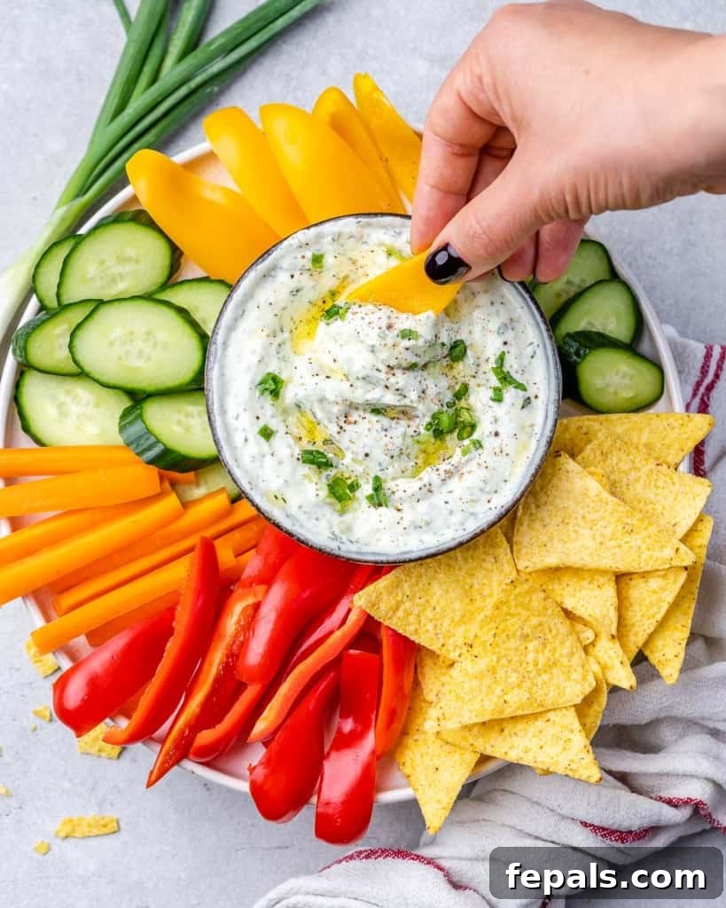 A hand holding crispy tortilla chips, poised to dip into a bowl of creamy herbed cottage cheese dip, surrounded by an abundant platter of fresh vegetables and additional chips.