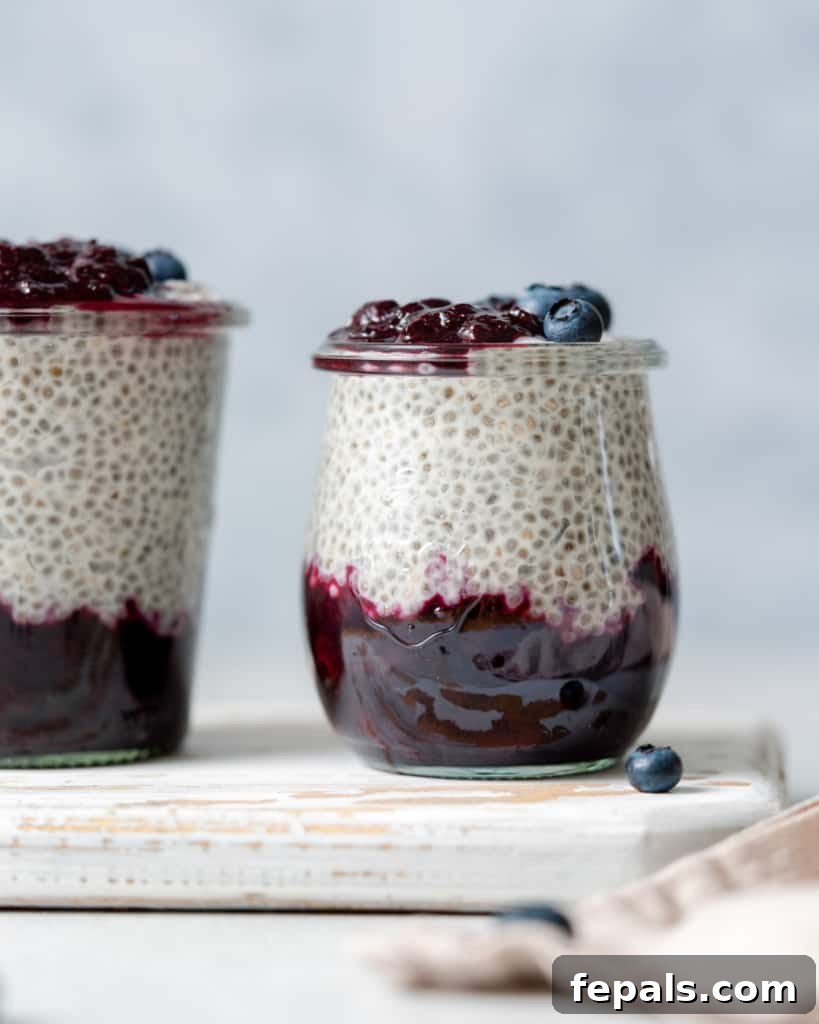 Side shot of a glass jar with blueberry jam at the bottom and chia pudding on the top layer, topped with more fresh blueberries and a spoon.