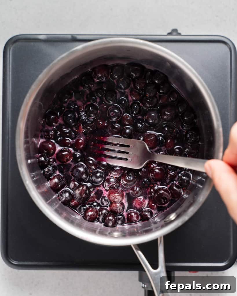 Mashing frozen blueberries with a fork in a saucepan, beginning to break down the fruit for sauce.