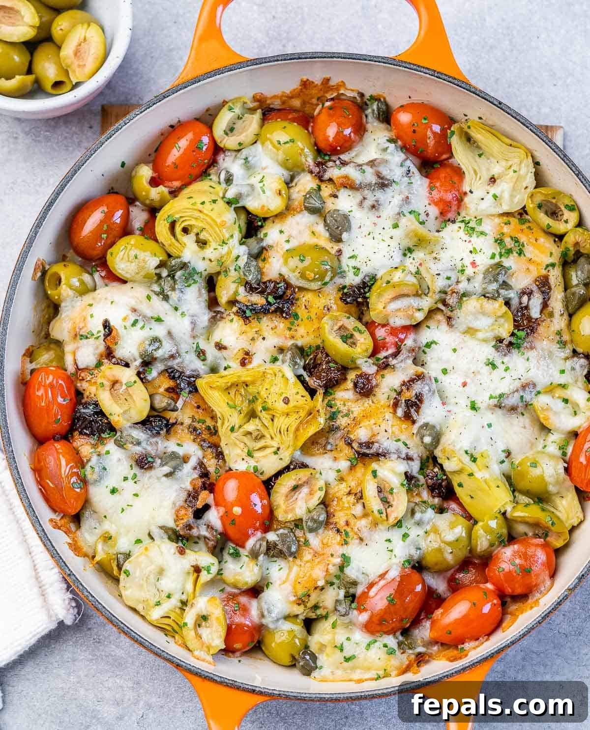 Baked Italian chicken in a skillet with tomatoes, olives, artichokes, capers and cheese, garnished with fresh herbs.
