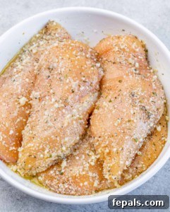 Raw chicken in a bowl covered with parmesan and seasonings.