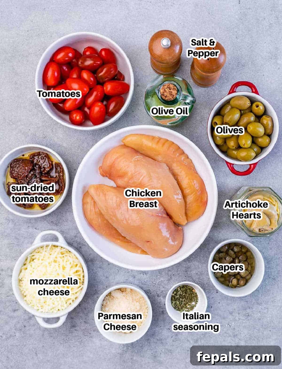 A flat lay of fresh ingredients laid out on a wooden board, including chicken breasts, cherry tomatoes, olives, artichoke hearts, sun-dried tomatoes, capers, mozzarella cheese, olive oil, and Italian seasonings.