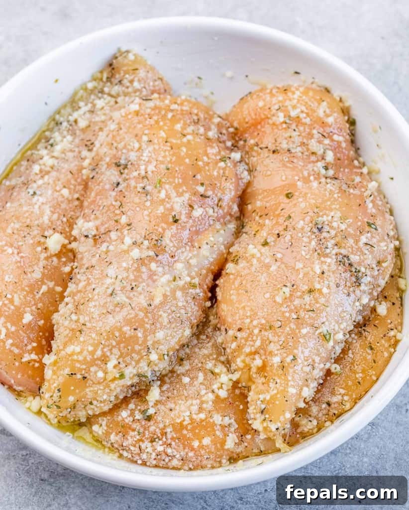 Raw chicken breasts in a bowl, coated with Italian seasonings, olive oil, and grated Parmesan cheese, ready for searing.