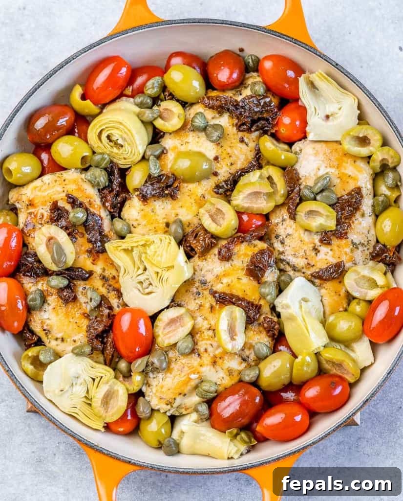Chicken in a skillet topped with a colorful mix of cherry tomatoes, artichoke hearts, and olives, before baking.