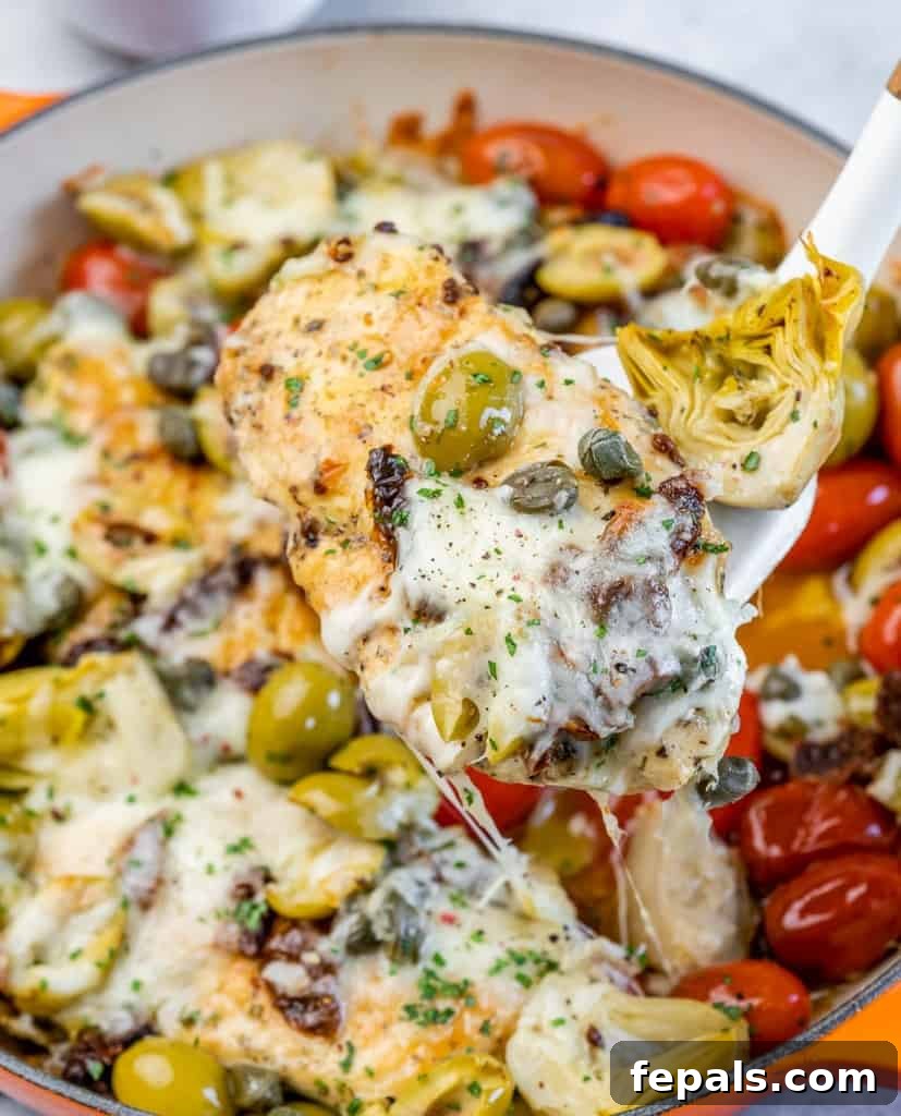 A spatula lifting a succulent piece of baked Italian chicken breast, topped with melted mozzarella and fresh olives, from the skillet.