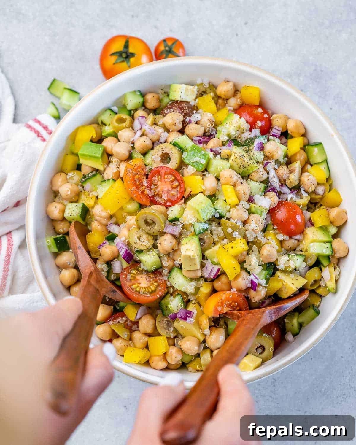 Mediterranean Chickpea Cucumber Quinoa Salad 3 A person tossing a vibrant chickpea quinoa salad with salad tongs in a large white bowl, highlighting its fresh ingredients.