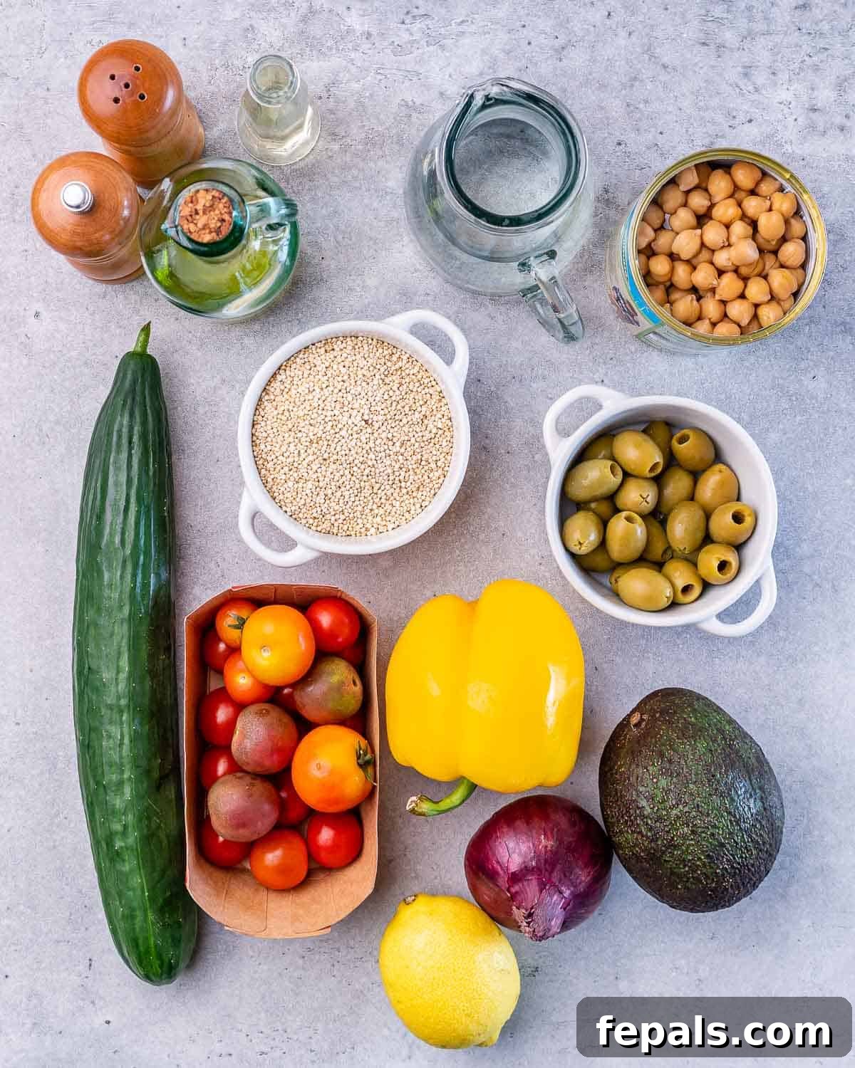 Mediterranean Chickpea Cucumber Quinoa Salad 4 A beautifully arranged flat lay of all fresh ingredients needed for the chickpea quinoa salad: uncooked quinoa, a cucumber, cherry tomatoes, an avocado, a red onion, a yellow bell pepper, a lemon, and a bowl of green olives.