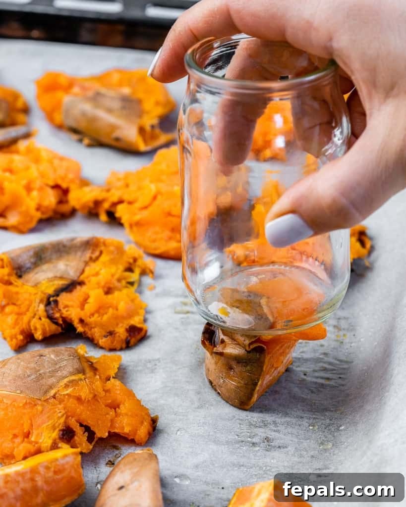 Golden Roasted Sweet Potato Smash 6 Hands using the bottom of a glass to gently smash a baked sweet potato piece on a parchment-lined baking sheet, demonstrating the smashing technique.