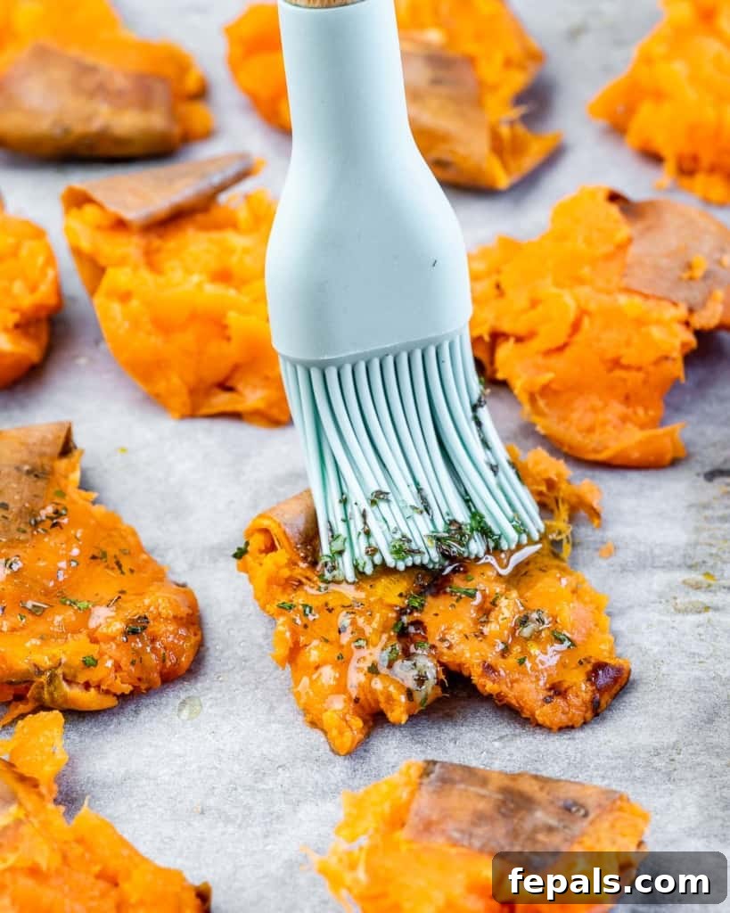Golden Roasted Sweet Potato Smash 7 A hand brushing the aromatic seasoning mix over a tray of freshly smashed sweet potatoes, preparing them for the final broiling step.