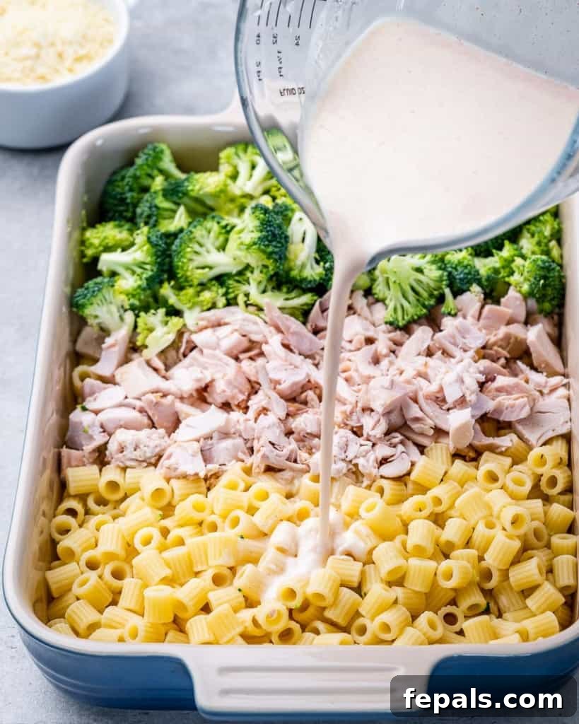 Pouring homemade Alfredo sauce over chicken, pasta and broccoli in a baking dish.