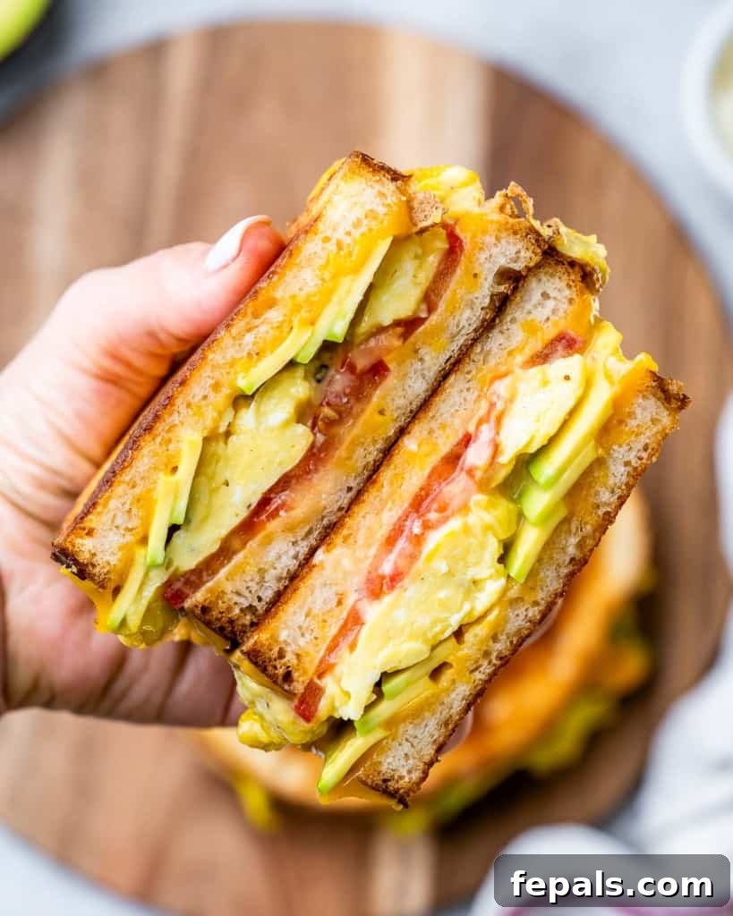 A hand holding a perfectly crafted breakfast egg sandwich, highlighting the layers of egg, cheese, avocado, and tomato.