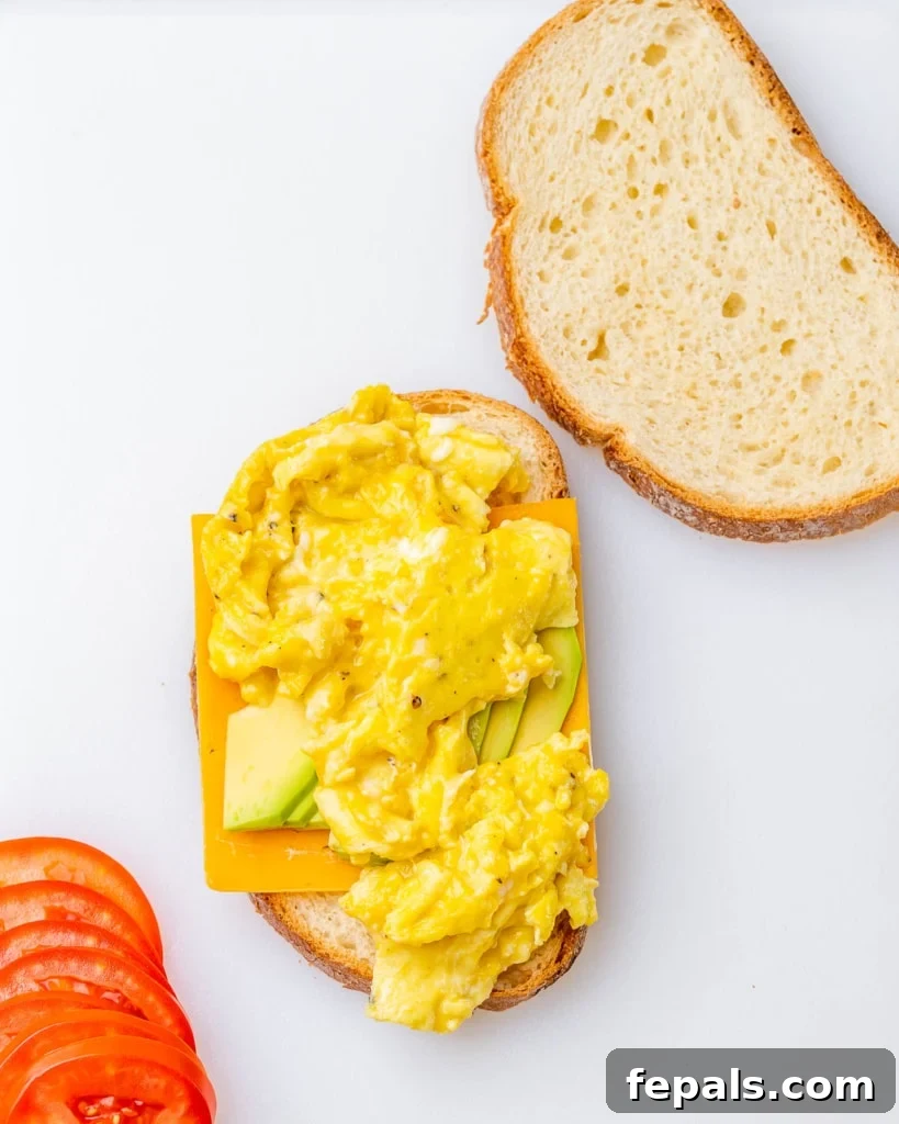 The assembly process of a breakfast egg sandwich, showing cheese, avocado, and scrambled eggs being layered onto a slice of bread.