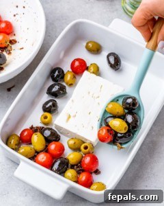 feta cheese block in a white dish with hand holding spoon and adding olives and tomato mixture