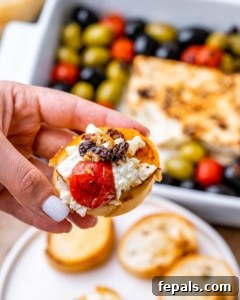 hand holding a baguette topped with feta cheese dip and baked tomatoes and olives