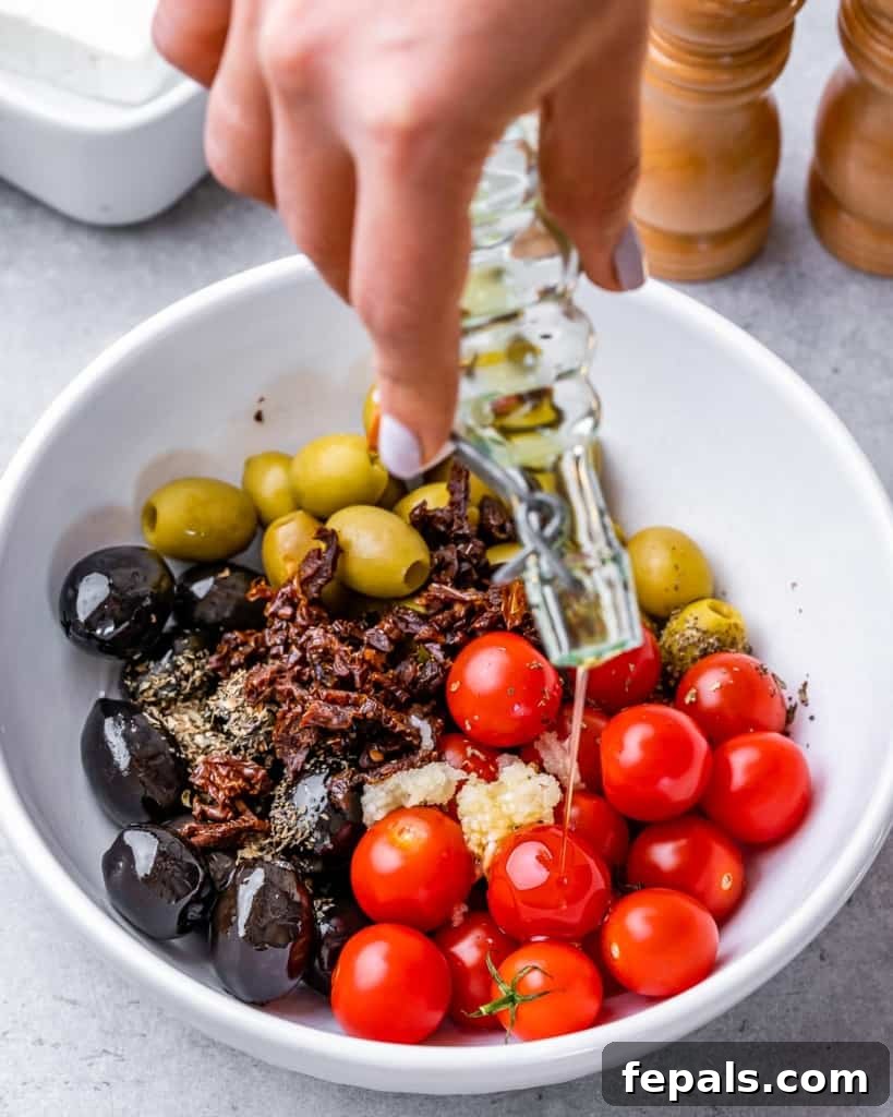 hand holding a jar of oil being poured over tomatoes and olives in a white bowl