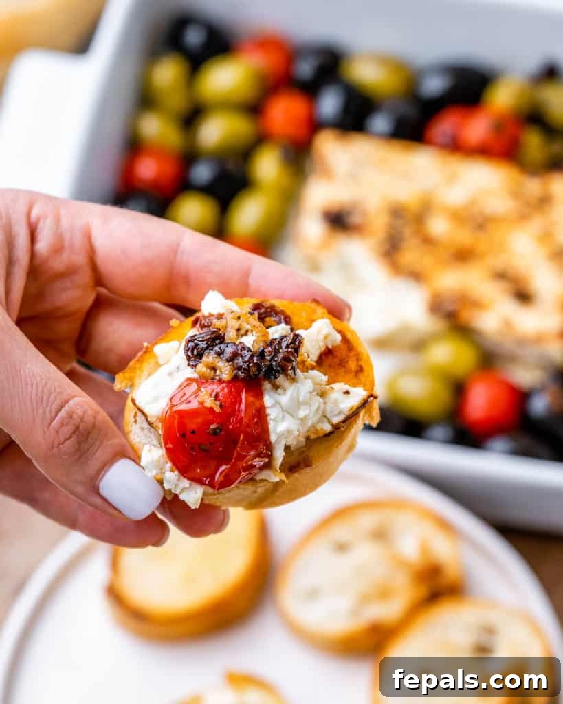 hand holding a baguette topped with feta cheese dip and baked tomatoes and olives, ready to eat