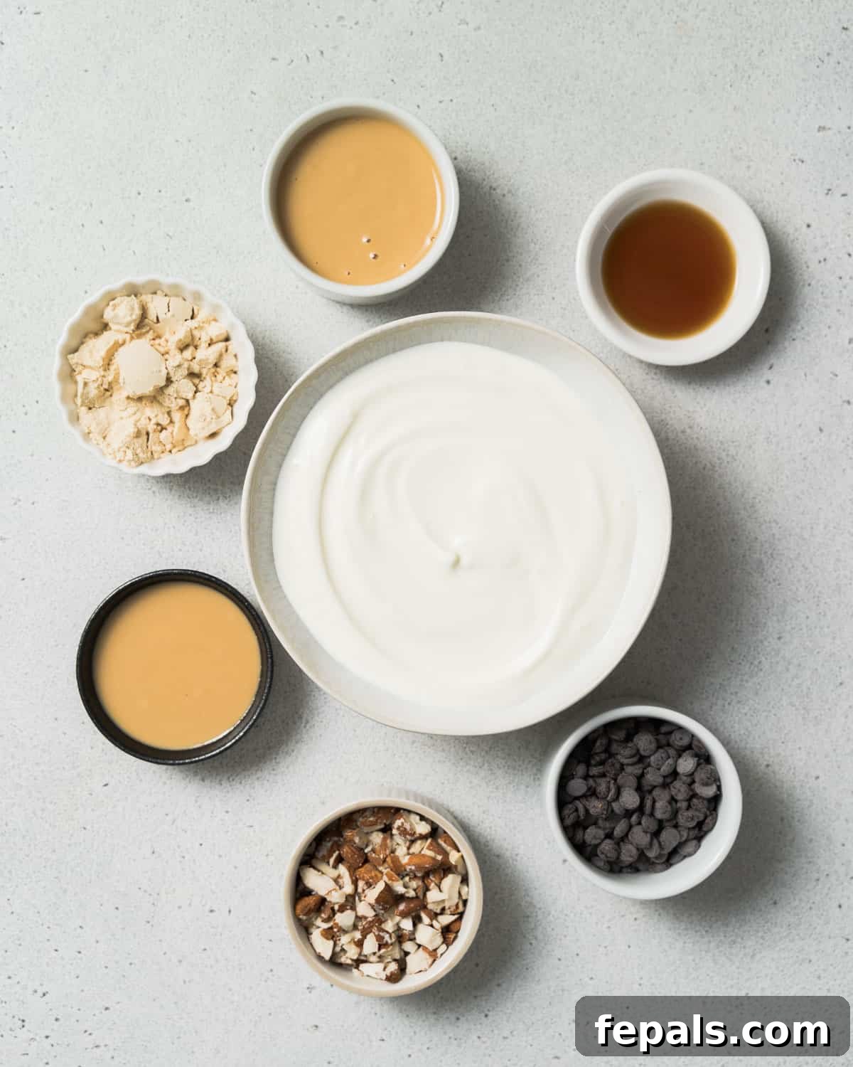 Peanut Butter Yogurt Frosty Bark 4 Bowls of yogurt, chocolate chips, almonds, peanut butter and maple syrup laid out, ready for preparation.