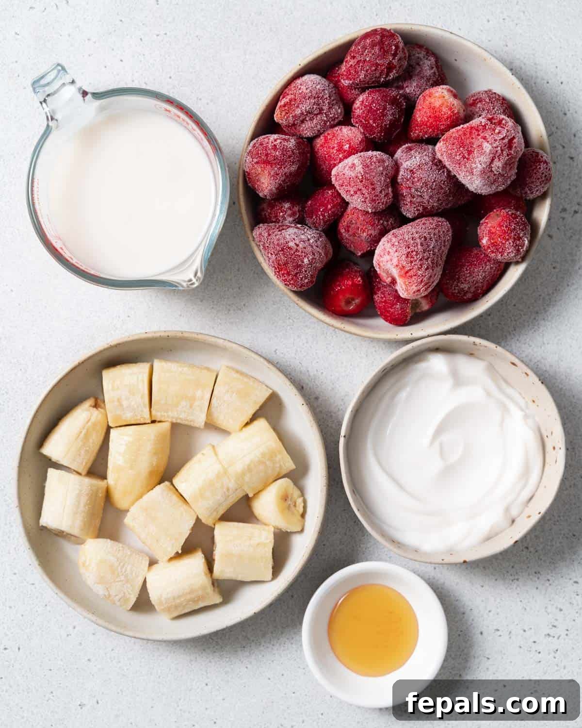 Frozen strawberries and banana slices in bowls with honey, Greek yogurt, and milk – all the ingredients laid out.