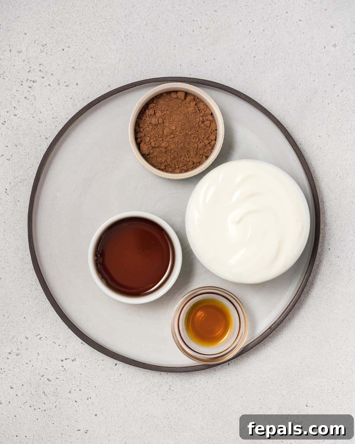Four small bowls containing Greek yogurt, cocoa powder, maple syrup, and vanilla extract, illustrating the simple ingredients needed.