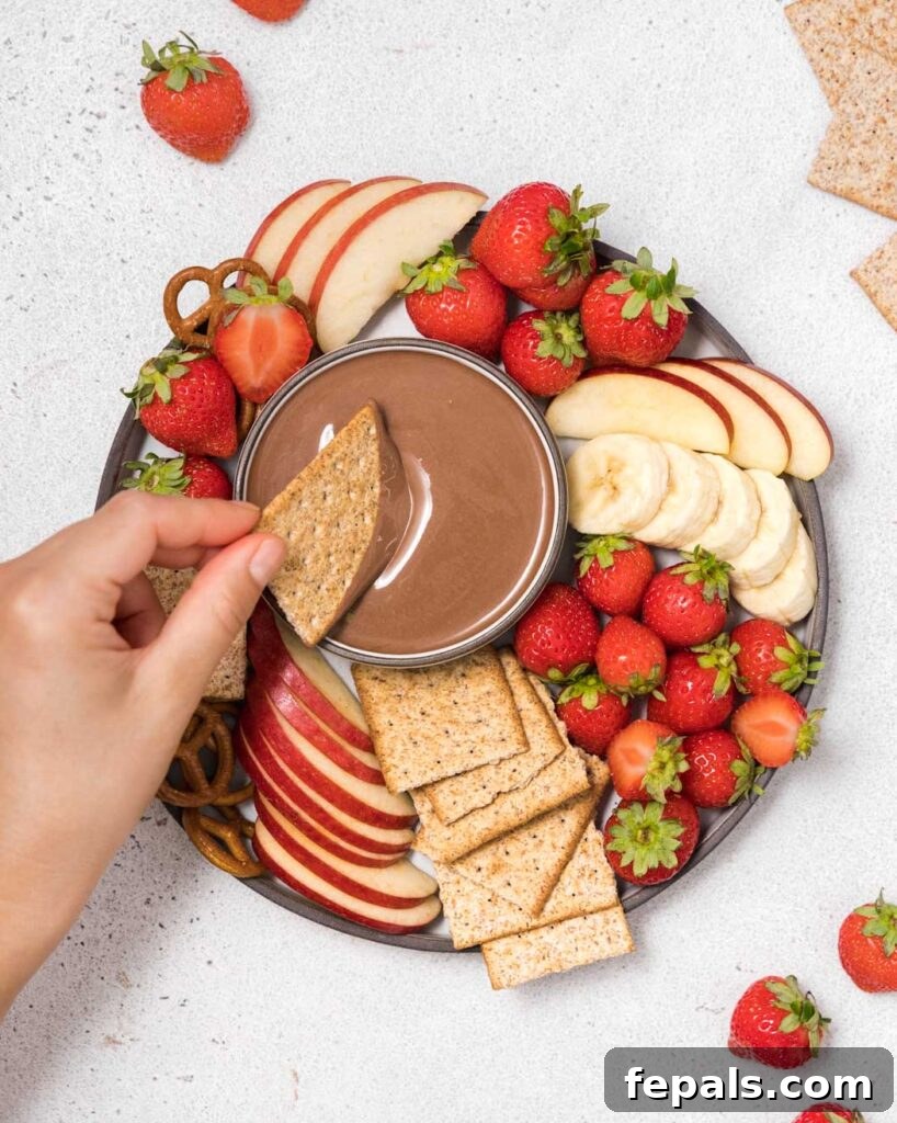 Hand reaching with a cracker into a bowl of chocolate dip, emphasizing its creamy texture and inviting nature.
