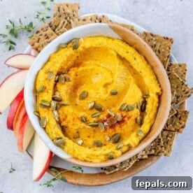 Top view of round white bowl with pumpkin hummus that's orange in color, topped with pumpkin seeds and a side of crackers by the bowl.