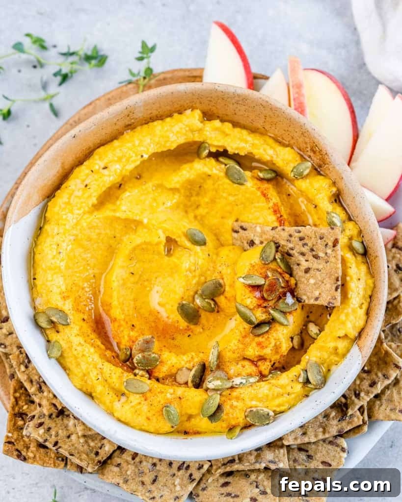 Spiced Pumpkin Hummus 9 top view or hummus thats orange in color and topped with pumpkin seeds in a round white bowl and a side of crackers