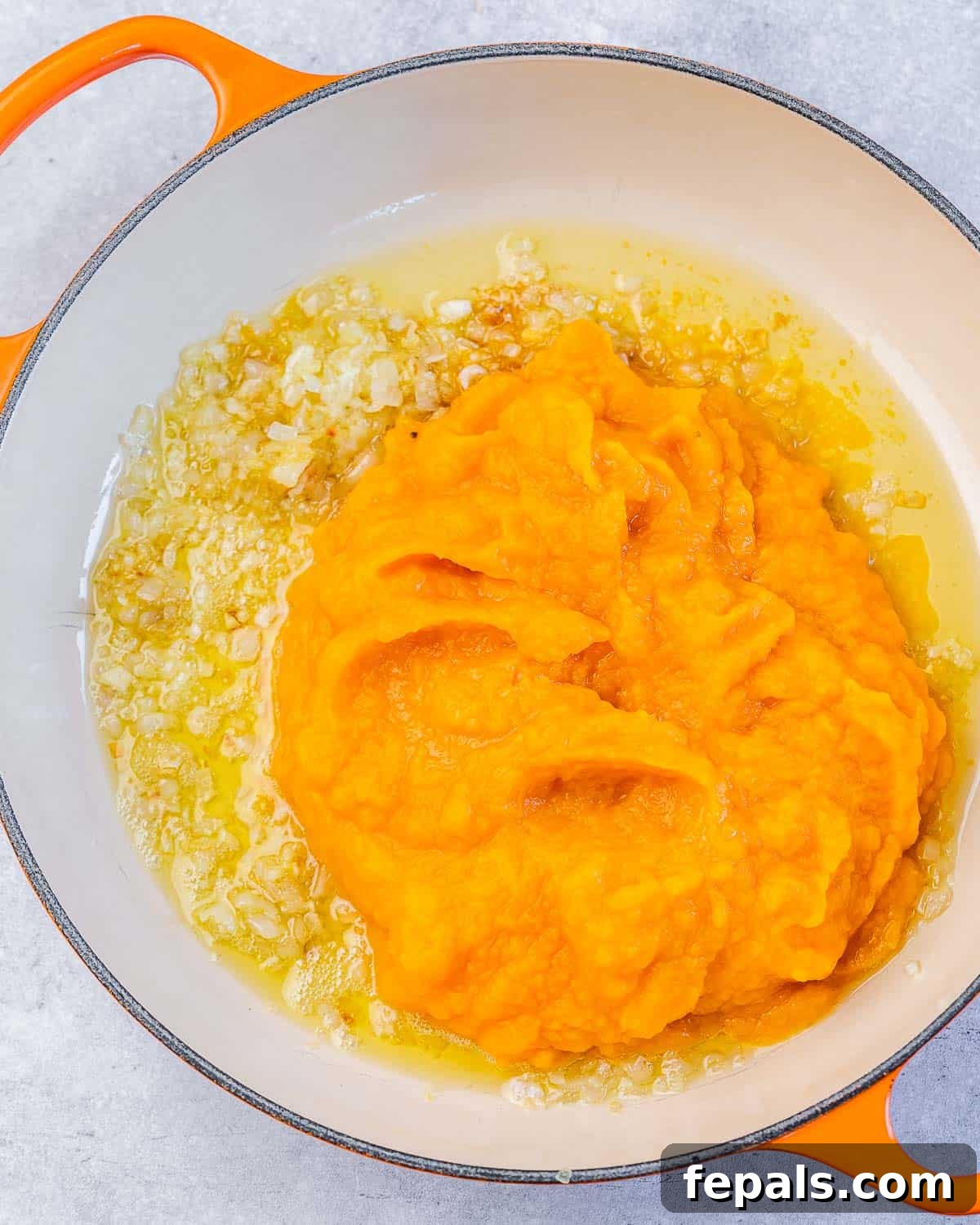 A hand adding pumpkin puree to sautéed shallots and garlic in a pan.