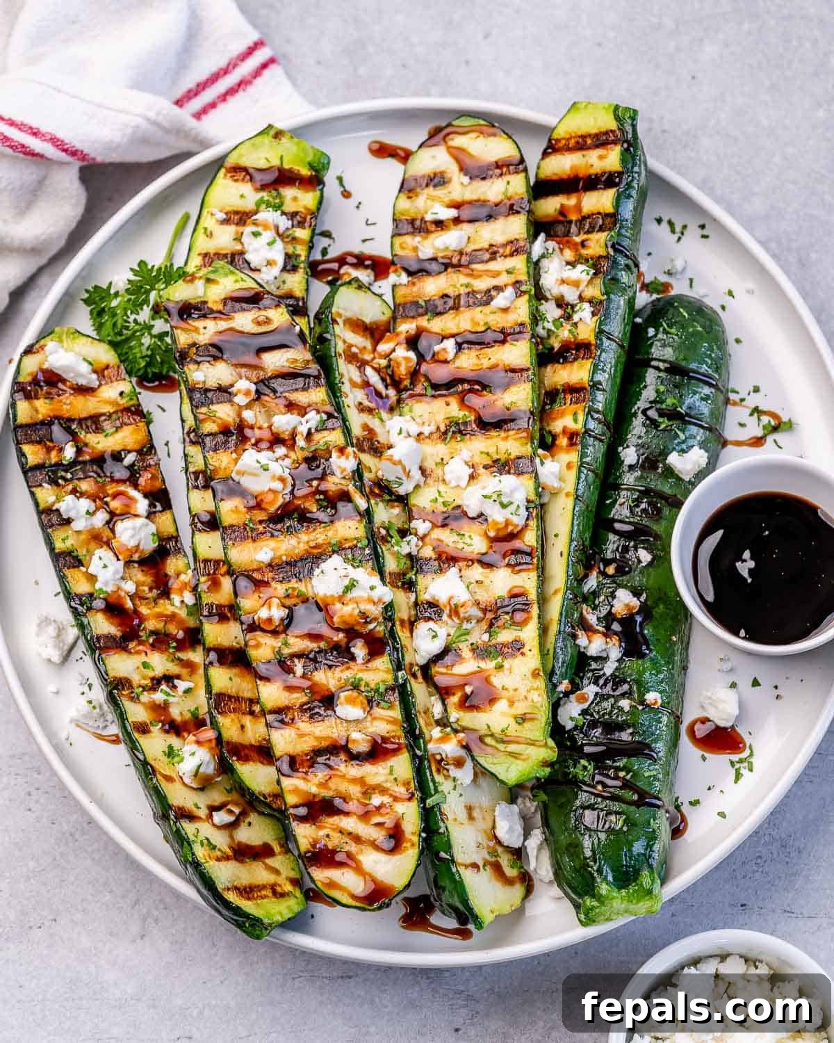 Top view of grilled zucchini halves on a round white plate drizzled with balsamic vinegar and goat cheese.