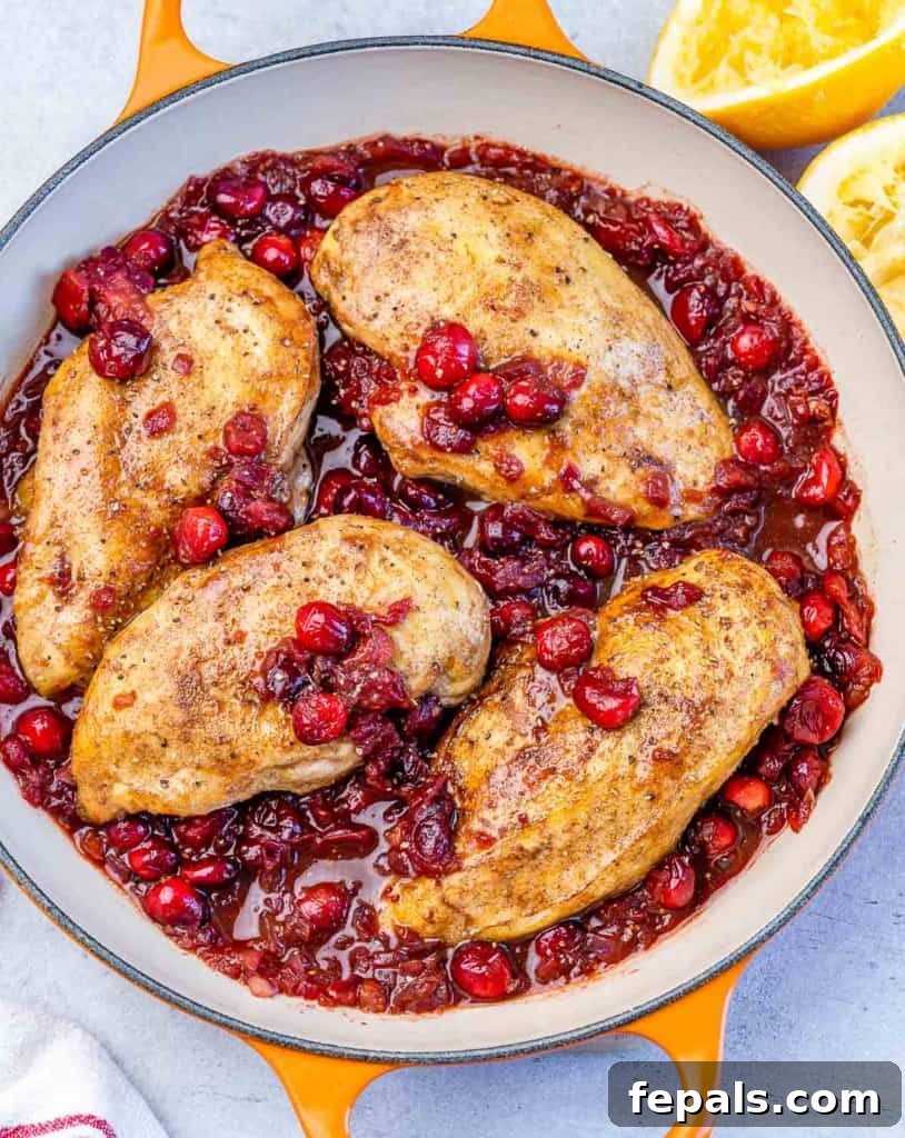 Top view of 4 baked chicken breasts nestled over a bed of vibrant cranberry sauce in an orange-colored skillet.