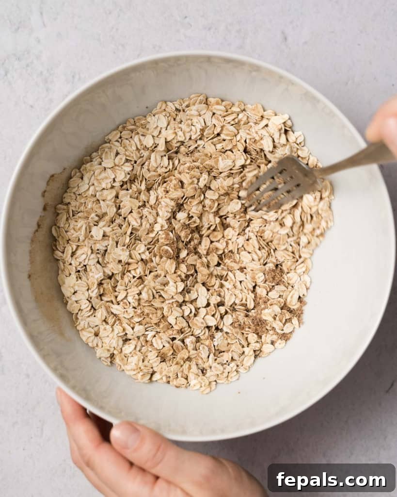 Spiced Pumpkin Power Morsels 6 A fork actively mixing rolled oats with a blend of spices in a large white bowl, ensuring all ingredients are evenly distributed.