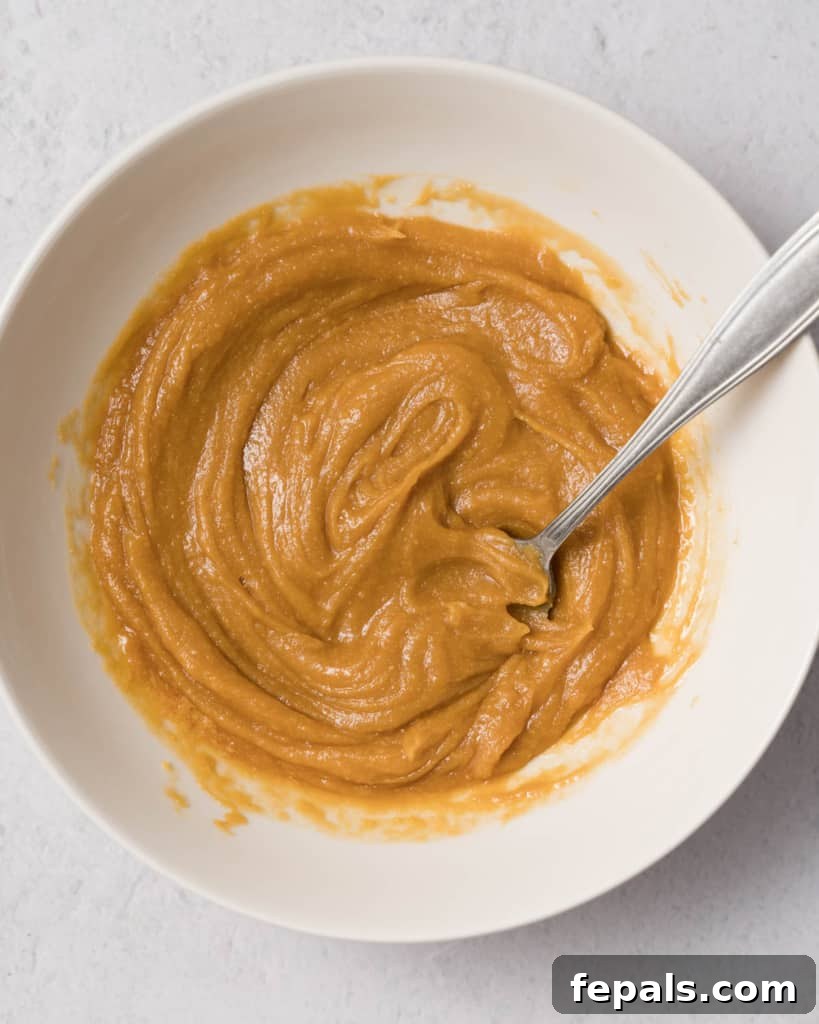 Spiced Pumpkin Power Morsels 7 A spoon stirring together creamy peanut butter, golden maple syrup, and vanilla extract in a white bowl, creating a smooth mixture.