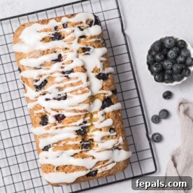 Zesty Blueberry Lemon Loaf 25 top view of a baked blueberry bread with icing drizzled on top on a wire wrack and a side of fresh blueberries in a small bowl