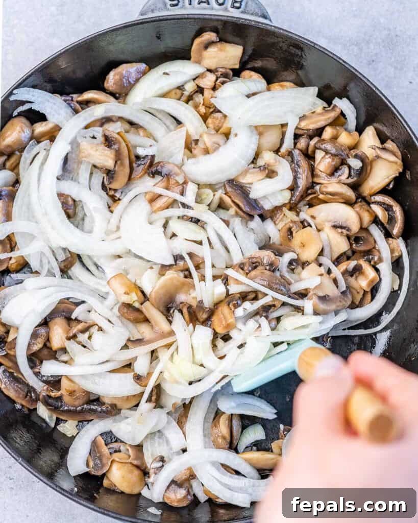 Thinly sliced onions are added to the sautéed mushrooms in a black skillet, ready to be cooked further for the casserole sauce.