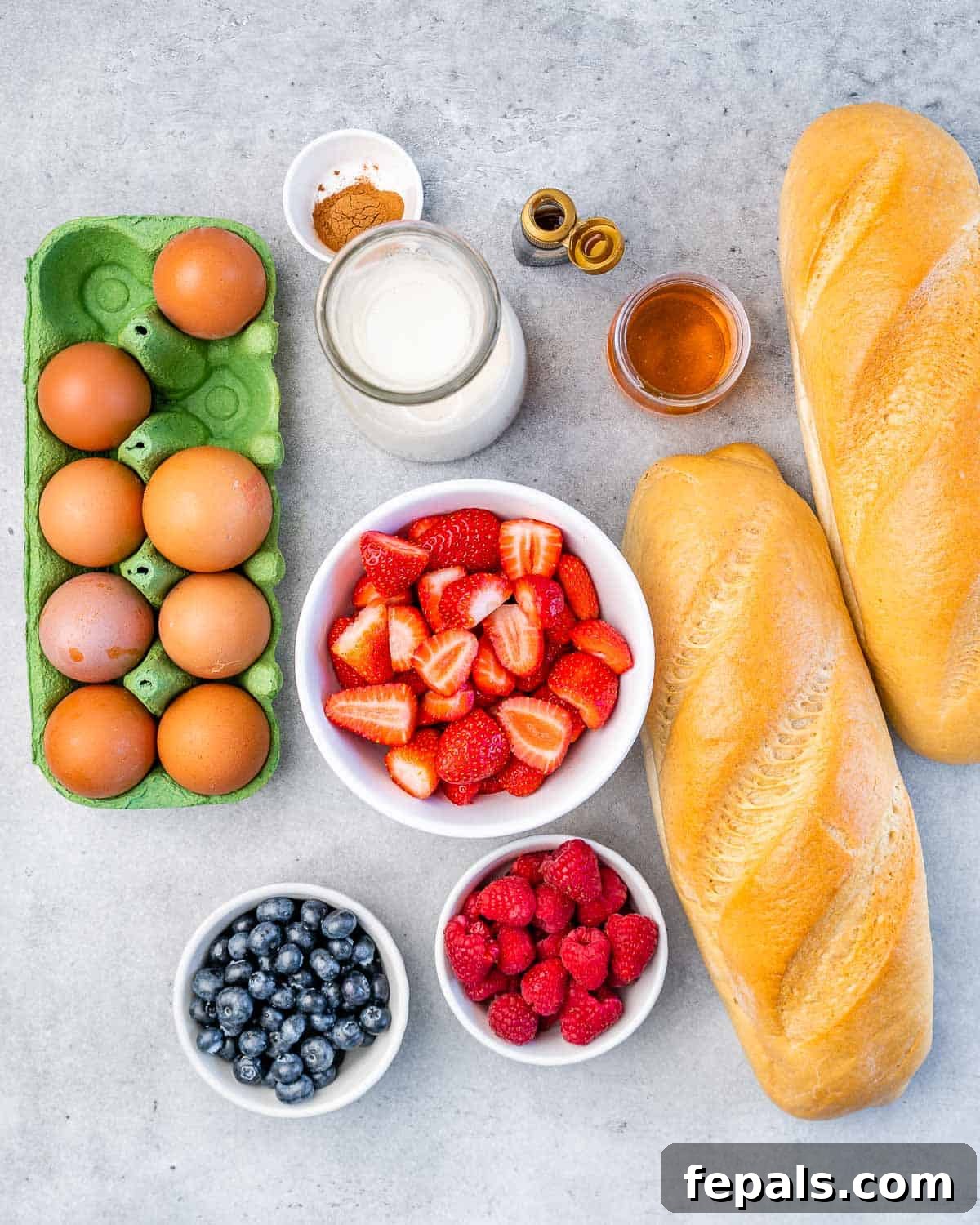 Two loaves of French bread, a bowl of mixed berries, eggs, milk, vanilla extract, cinnamon, and honey, all portioned for the recipe.