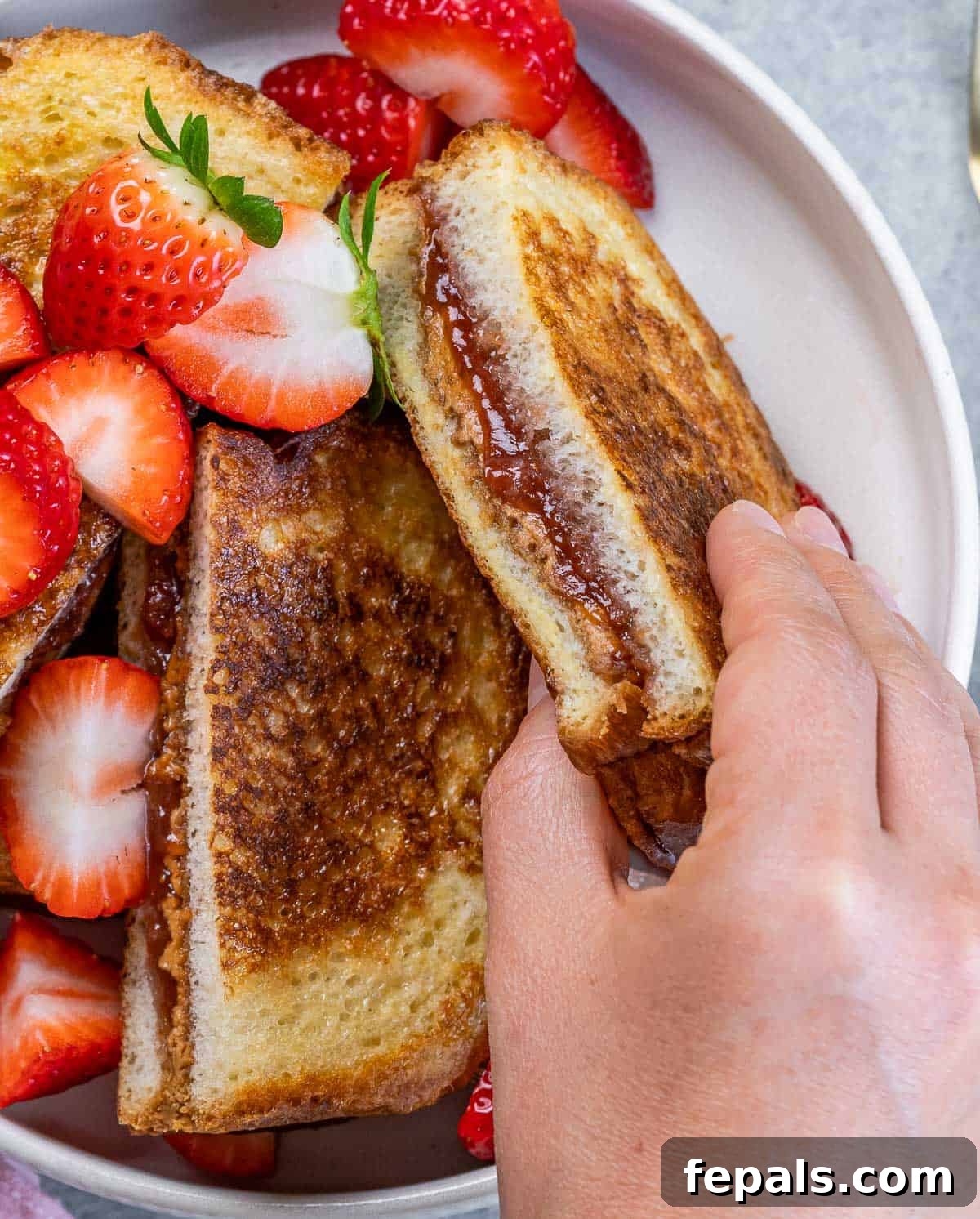 Peanut Butter Jelly French Toast Surprise 10 Hand picking up a peanut butter jelly French toast near sliced strawberries.