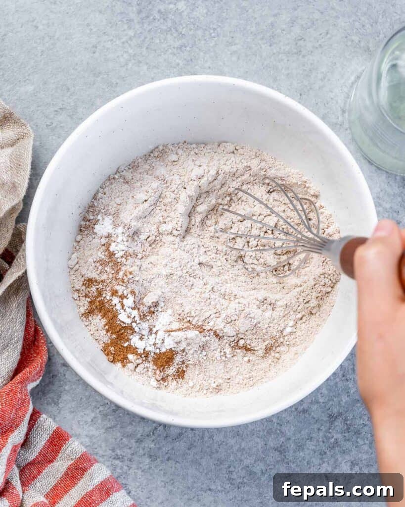 A whisk blending whole wheat flour with cinnamon and baking powder in a white bowl, showing the initial dry ingredients being combined.