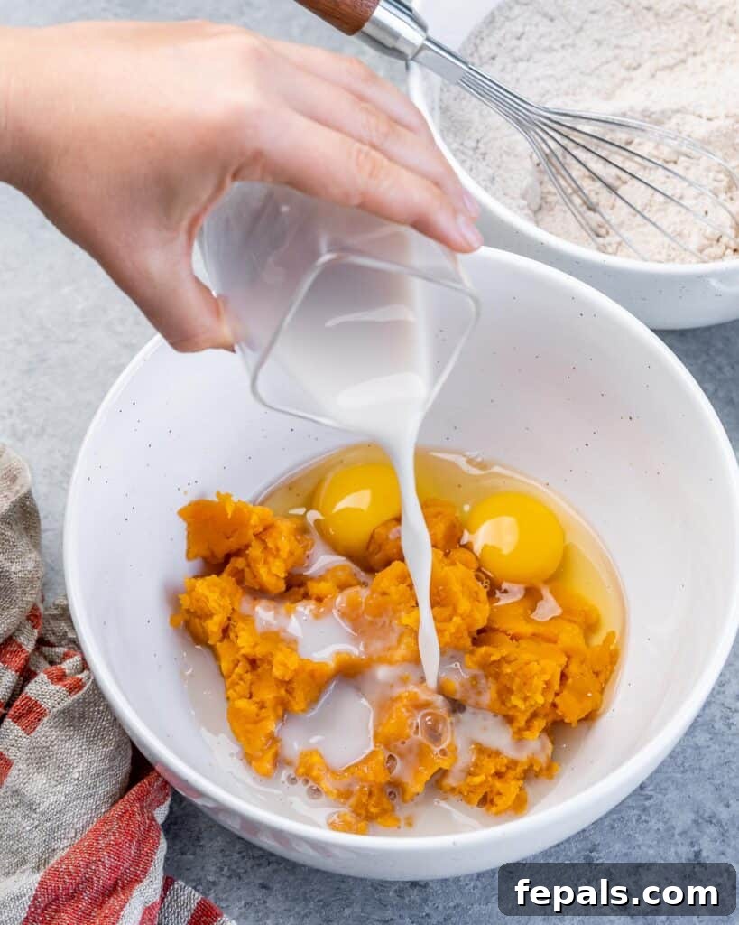 Pouring milk into a bowl containing mashed sweet potatoes and eggs, demonstrating the integration of liquids into the wet mixture.
