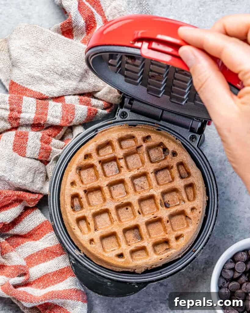 A hand opening the waffle maker to reveal freshly made chocolate waffles, still steaming inside.