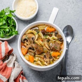 Hearty Beef Noodle Comfort 11 top view of a white bowl with beef noodle soup next to a small bowl of fresh parmesan cheese and fresh parsley