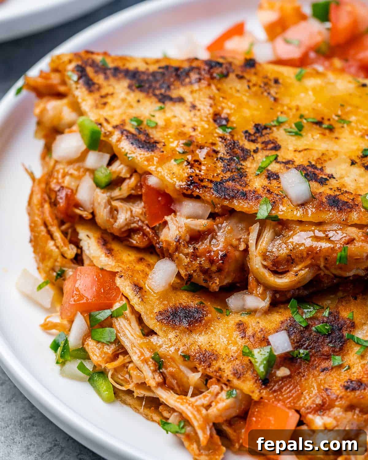Crispy Birria Chicken Dipping Tacos 3 Bir tabakta istiflenmiş tavuk Birria tacosunun yakın çekim görünümü, çıtır tortillaları ve erimiş peyniri gösteriyor.