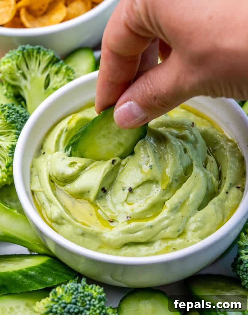 hand holding a peace of cucumber and dipping in the avocado rand dip in a bowl 