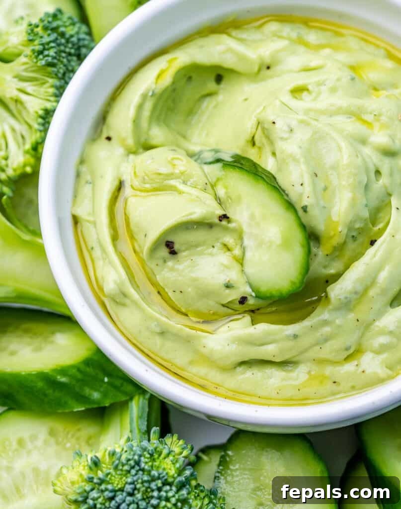 close up green looking avocado ranch dip in a white bowl with a slice of cucumber in the bowl