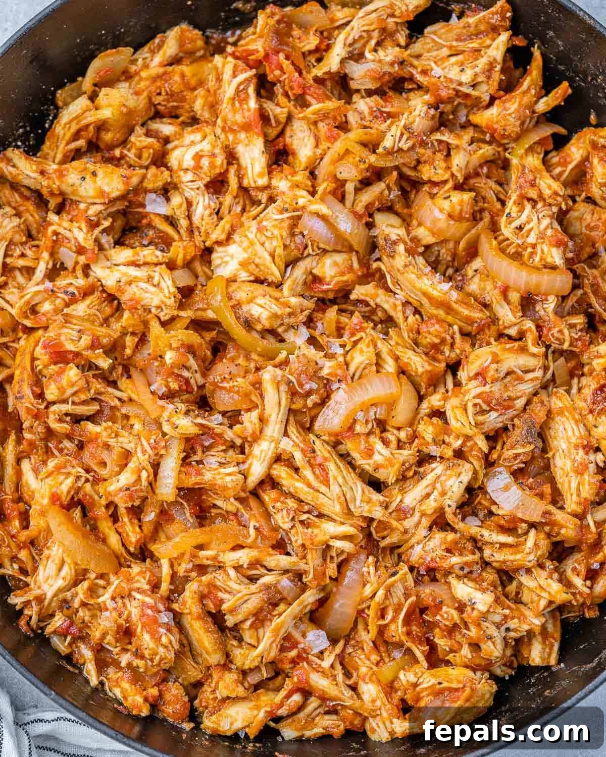 The Ultimate Chicken Tinga 7 A close-up shot of Chicken Tinga cooking in a large skillet, with the vibrant red-orange sauce bubbling gently around the tender shredded chicken pieces.