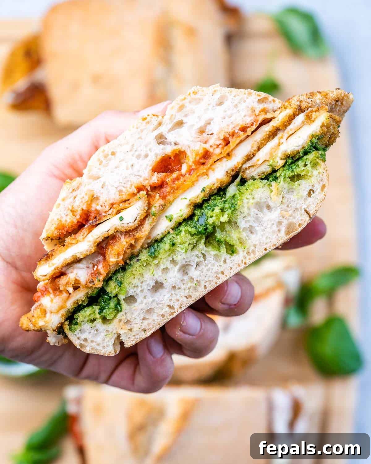 Crispy Chicken Parmesan Sandwich Outshines Takeout 2 A person holding a delicious Chicken Parmesan Sandwich with a visible crispy breaded chicken, pesto, marinara sauce, and melted mozzarella between toasted ciabatta bread.