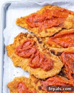 Close-up of crispy golden chicken cutlets topped with dollops of marinara sauce on a baking tray.