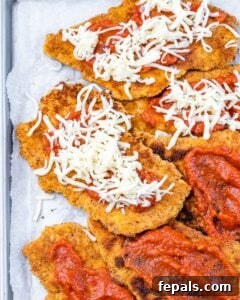 Breaded chicken cutlets on a baking sheet, topped with marinara sauce and shredded mozzarella, ready for the oven.