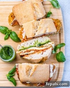 Sliced Chicken Parmesan Sandwiches served on a wooden board with pesto sauce and fresh basil scattered around.
